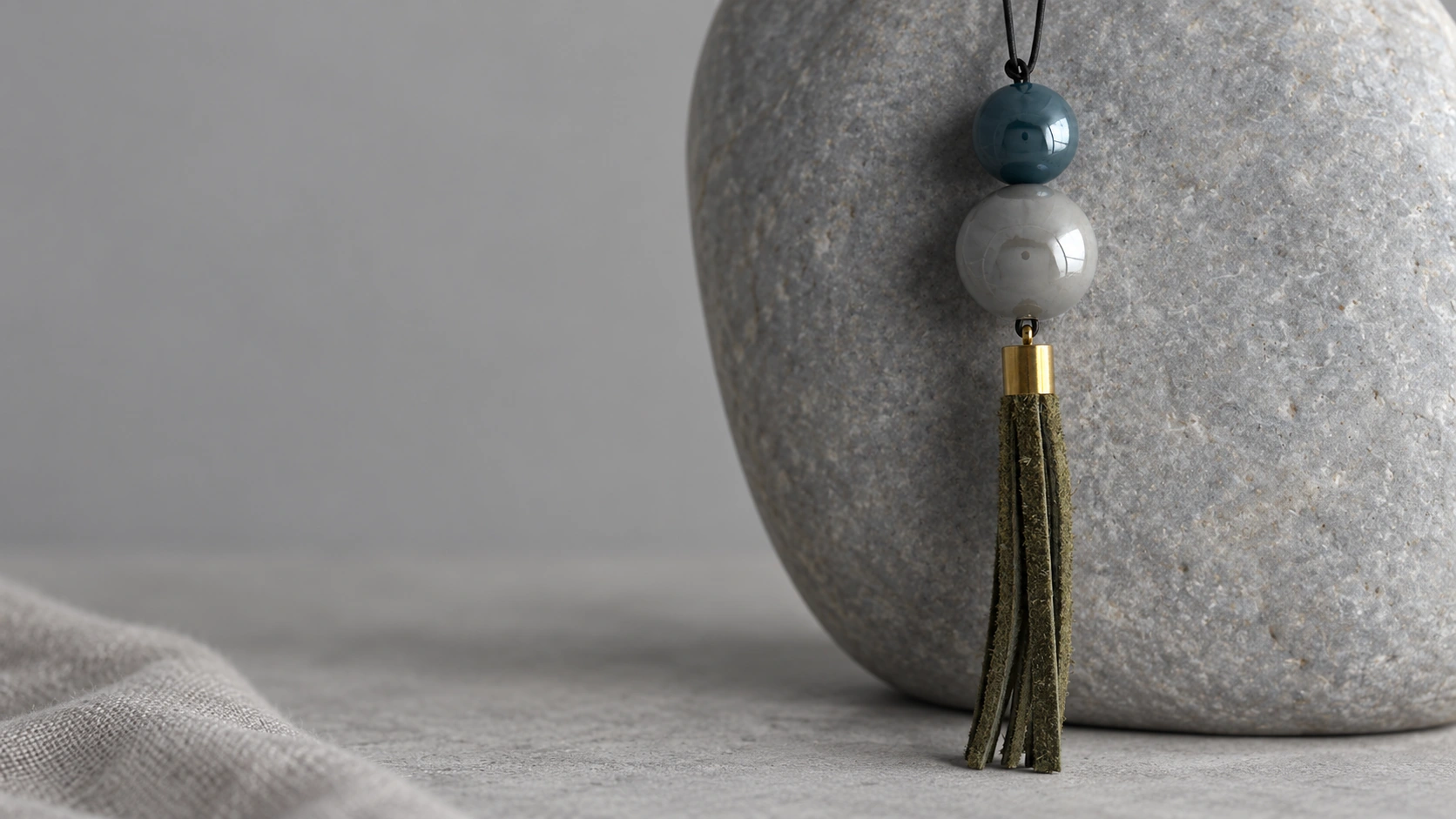 TERRA ceramic pendant with an olive leather tassel against a smooth grey river stone